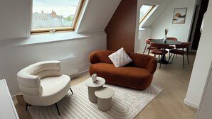Un salon moderne avec un fauteuil blanc aux lignes courbes, un canapé marron agrémenté d’un coussin blanc et une petite table ronde. On aperçoit à l’arrière-plan un coin repas avec une table et des chaises.