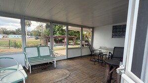 Un espace extérieur abrité, doté d’un sol en briques, de chaises vertes et blanches, d’une table et offrant une vue sur un parc avec un kiosque.