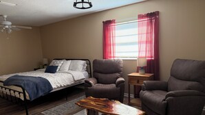 Une chambre à coucher avec un lit, deux fauteuils, une table basse en bois, un ventilateur de plafond et une fenêtre avec des rideaux rouges.