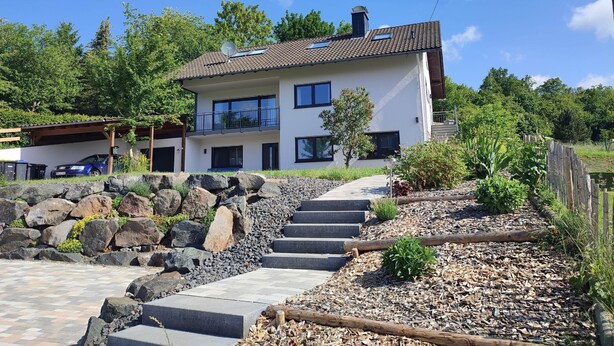 A two-story house with a stone pathway leading to the entrance, surrounded by greenery and a wooden fence. - Vacation home Franzi with barbecue, garden and terrace, quiet location on the outskirts up to 6 pers (Grebenhain)