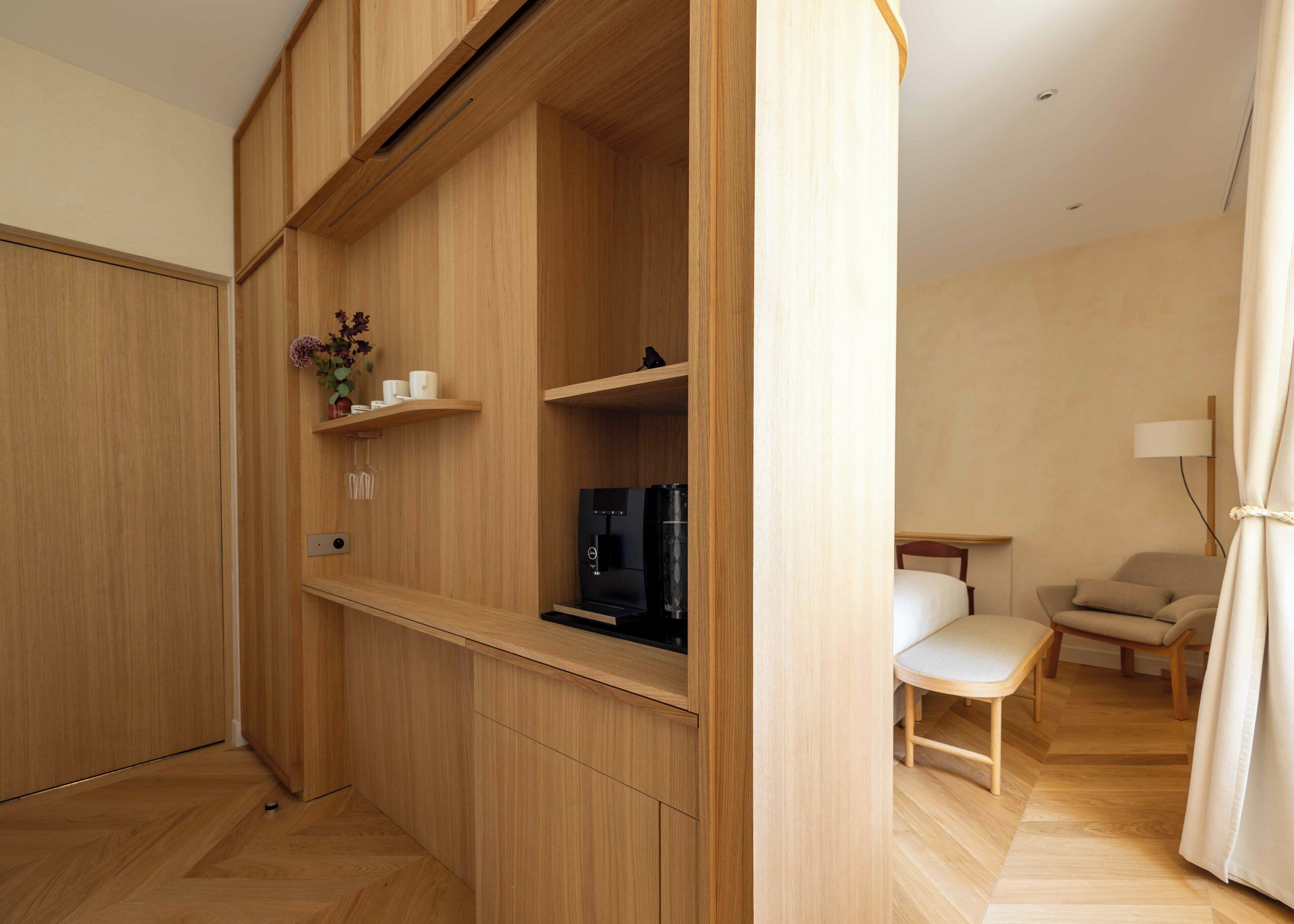 A modern, compact room with built-in wooden cabinets, a coffee machine, and a small seating area with a chair and table.