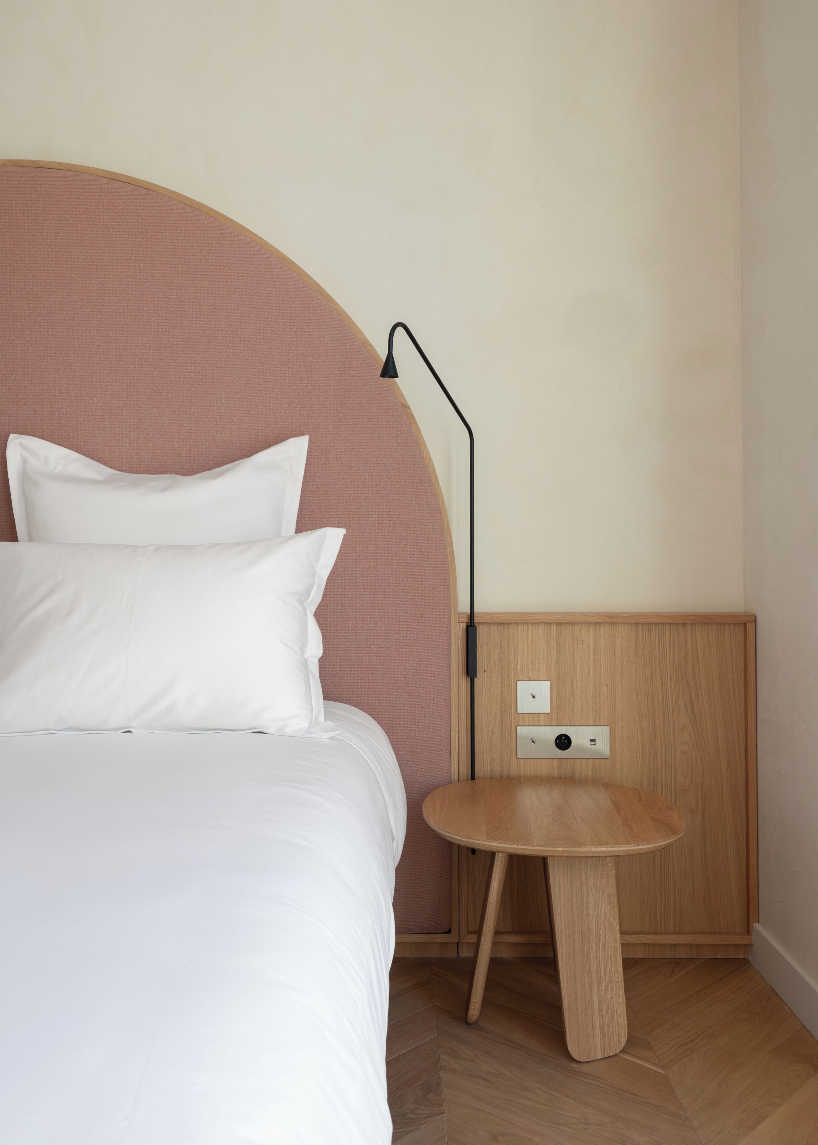 Una cama bien hecha con sábanas y almohadas blancas, una mesita de noche redonda de madera con una lámpara, y un tablero de interruptores en la pared.