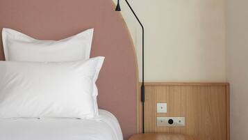 Una cama bien hecha con sábanas y almohadas blancas, una mesita de noche redonda de madera con una lámpara, y un tablero de interruptores en la pared.