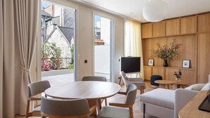 A modern living room with a wooden dining table, grey upholstered chairs, a sofa, and a TV. - Le N°3 par Champagne Thiénot (Reims)