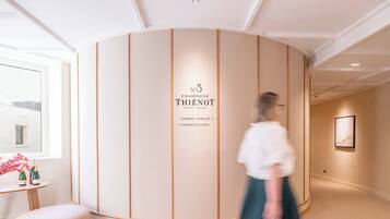 A hotel corridor with a curved wall displaying the name "Chambre Thienot" and a sign indicating room types. A woman in a white blouse and green skirt walks through the corridor.
