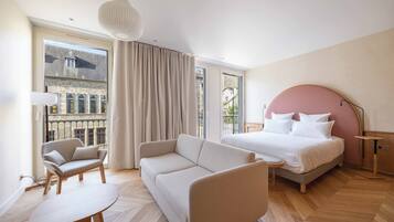 A modern hotel room with a large bed, a sofa, a small table, and a view of the city through large windows.