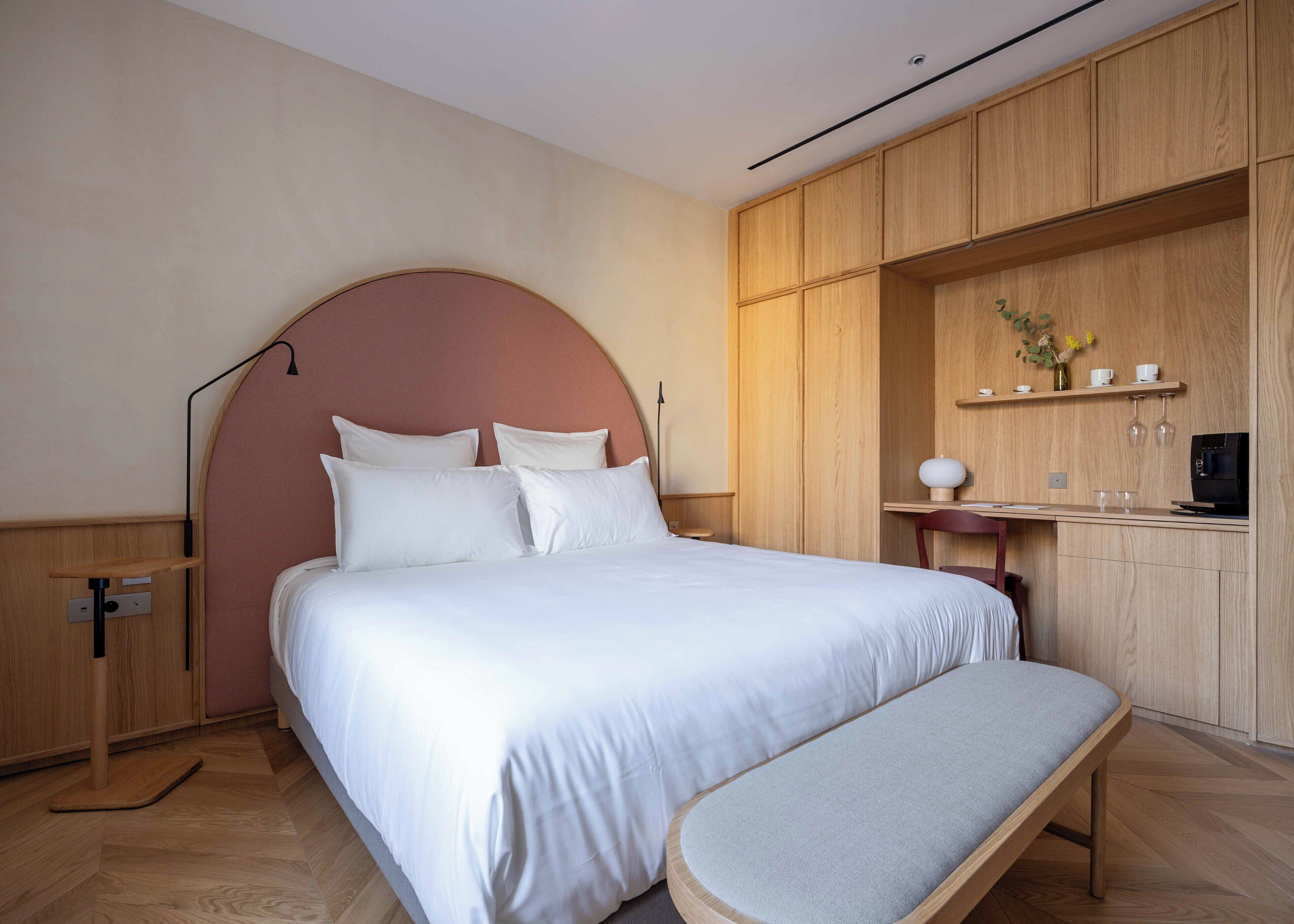 A modern bedroom with a large bed, a bench, wooden cabinets, and a small kitchenette area.