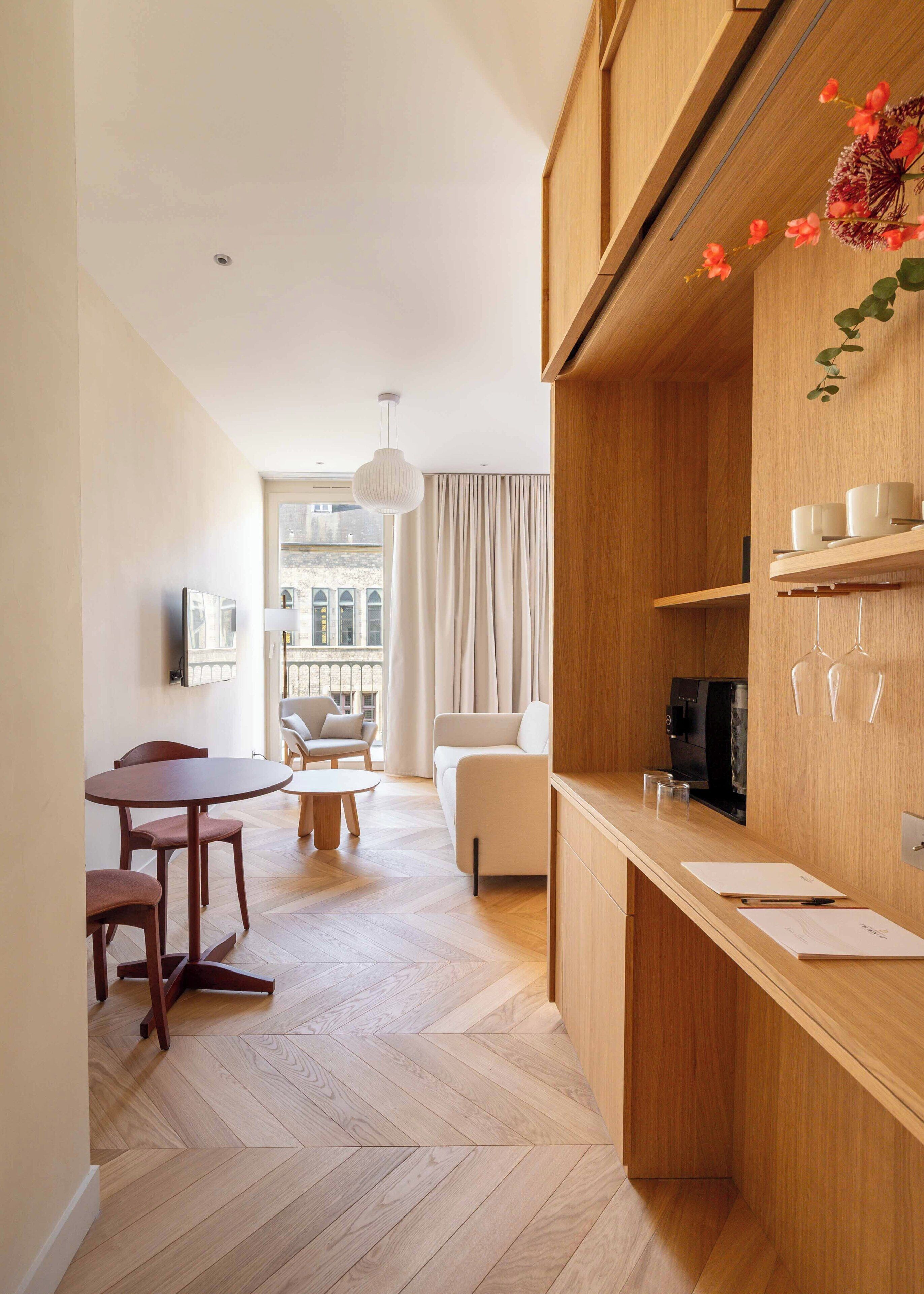 A modern kitchen with wooden cabinets, a dining area with a round table and chairs, and a view of a cityscape through large windows.