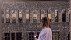 A person holding a glass of wine, overlooking a building with arched windows and a brick facade. - Le N°3 par Champagne Thiénot (Reims)