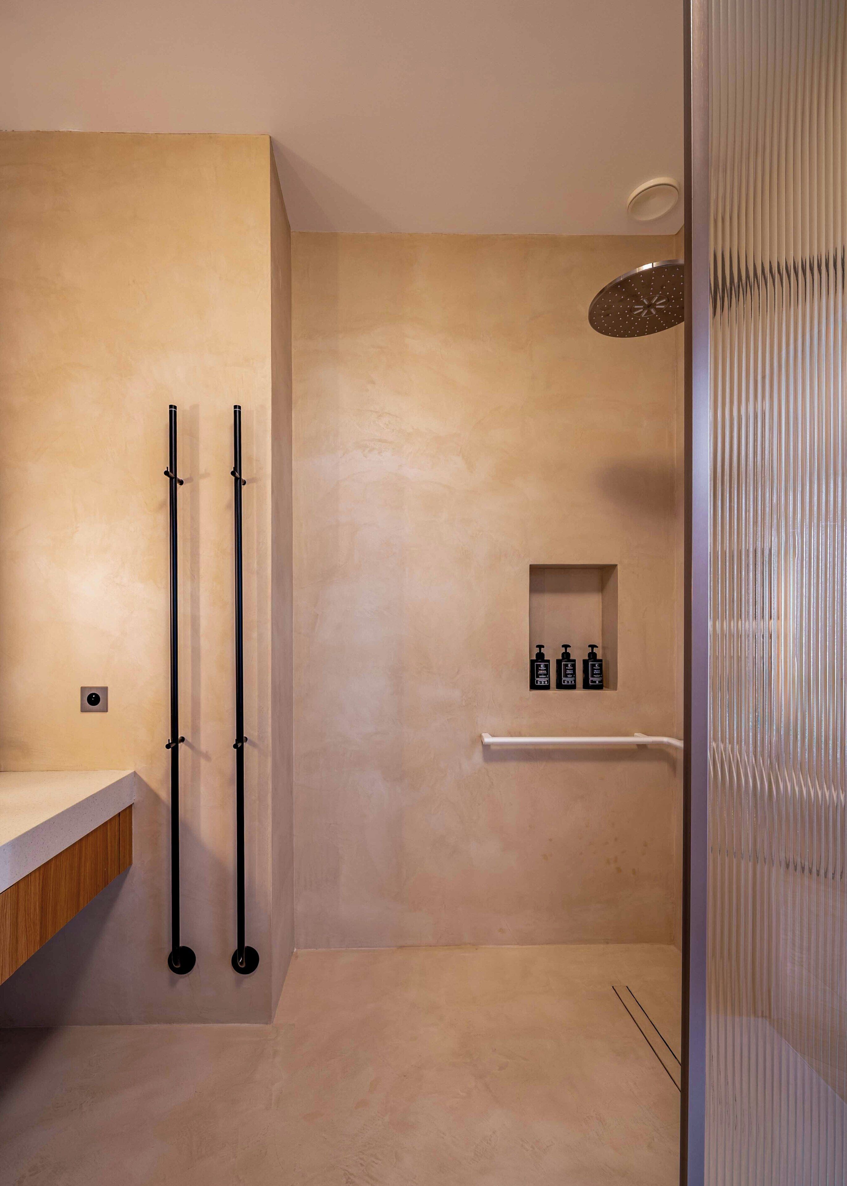Un baño moderno con una pared texturizada color beige, un mueble de tocador de madera, dos toalleros negros, un barra para toallas blanca y una pequeña estantería con artículos de aseo.