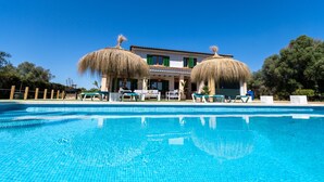 Une piscine avec des chaises longues, un bâtiment surmonté de deux parasols en chaume, et un ciel d’un bleu limpide.