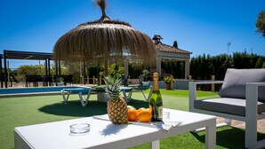 Un espace aménagé au bord de la piscine, avec une table dressée avec des fruits, une bouteille et des verres, une structure à toit de chaume et du mobilier d’extérieur.