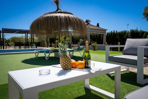 Un área junto a la piscina con una mesa dispuesta con frutas, una botella y vasos, una estructura de techo de paja y mobiliario exterior.