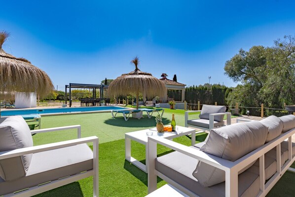 A poolside area with a thatched-roof gazebo, outdoor seating, and a clear blue sky.