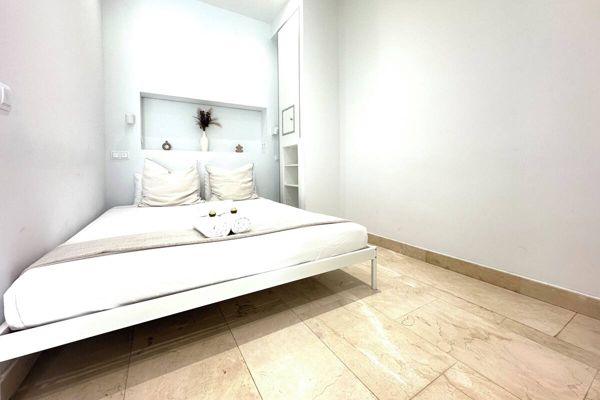 A minimalist bedroom with a single bed, a small bedside table, and a wall-mounted shelf with a vase.