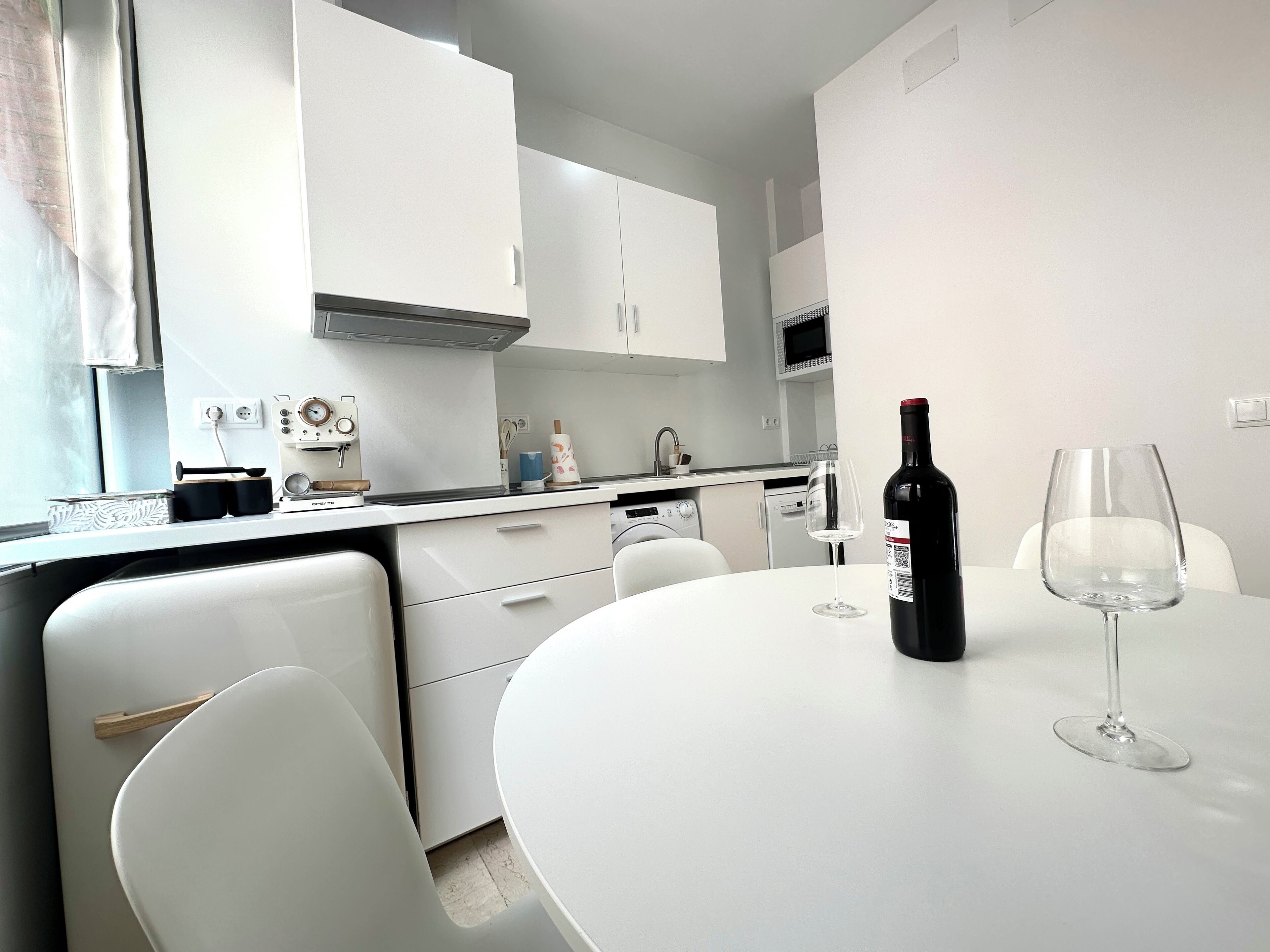 Una cocina moderna con armarios blancos, una mesa de comedor con dos sillas, una botella de vino y una copa de vino.