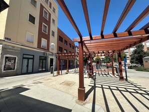 Un espacio al aire libre cubierto con una estructura de pérgola, un edificio con múltiples ventanas y un cielo azul despejado.