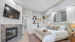 A modern bedroom with a fireplace, a large bed, a TV, and decorative shelves. - 7BR 5BA w Pool, Hot Tub, Sport Court & Game Room (DeSoto)