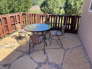 Une terrasse extérieure avec une table ronde et quatre chaises en osier, entourée d’une clôture en bois et de carreaux de pierre.