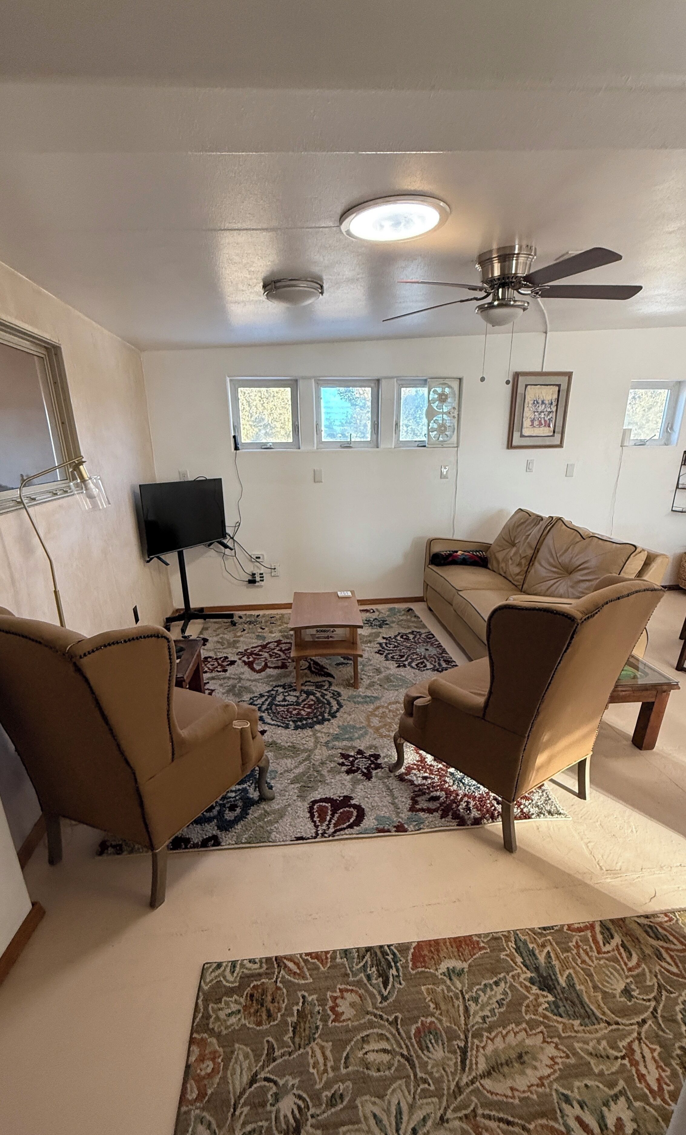 A living room with a ceiling fan, a TV, a sofa, two armchairs, and a patterned rug.