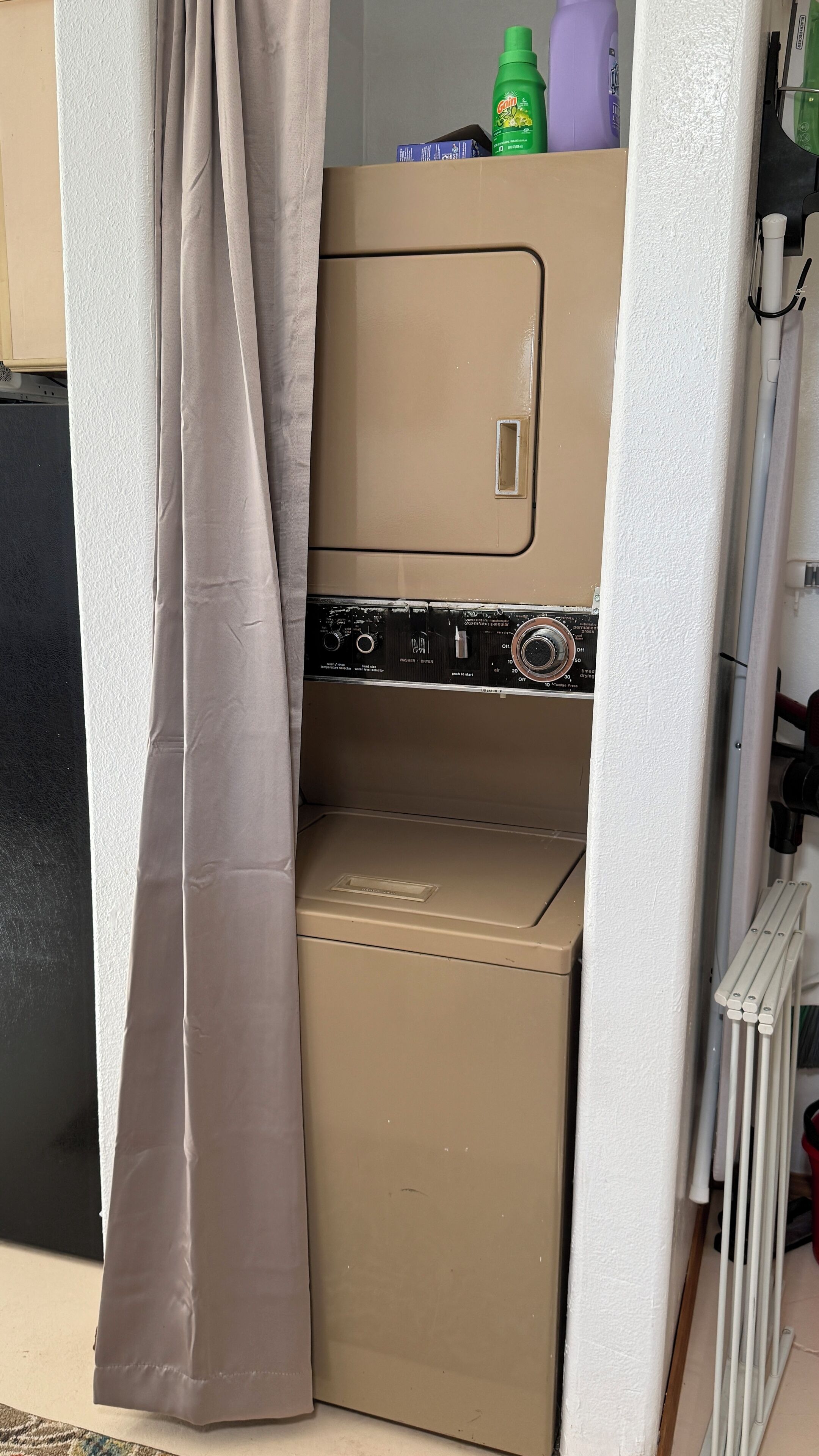 A compact laundry room with a washing machine and dryer, a curtain, and a shelf with cleaning supplies.