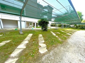 Un espace extérieur abrité, avec un chemin menant à l’entrée d’un bâtiment, un petit arbre et une clôture en métal.