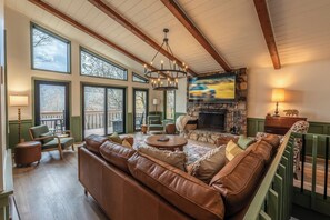 A spacious living room with a large brown leather sofa, a stone fireplace, and a television mounted on the wall. - Spacious 5-bdrm house in tranquil 7 Devils. Hottub & AC. < 1 mile to Hawksnest (Seven Devils)