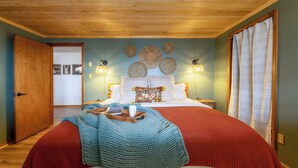 A bedroom with a wooden ceiling, a bed with red and white bedding, a blue knitted blanket, and wall decor. - Spacious 5-bdrm house in tranquil 7 Devils. Hottub & AC. < 1 mile to Hawksnest (Seven Devils)