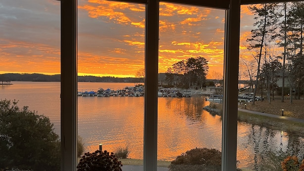 Un amplio ventanal con vista a un atardecer sobre un lago, barcos amarrados y una pequeña planta en maceta en el alféizar.