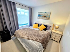 A bedroom with a bed, a nightstand, a lamp, a painting, and a window with curtains.