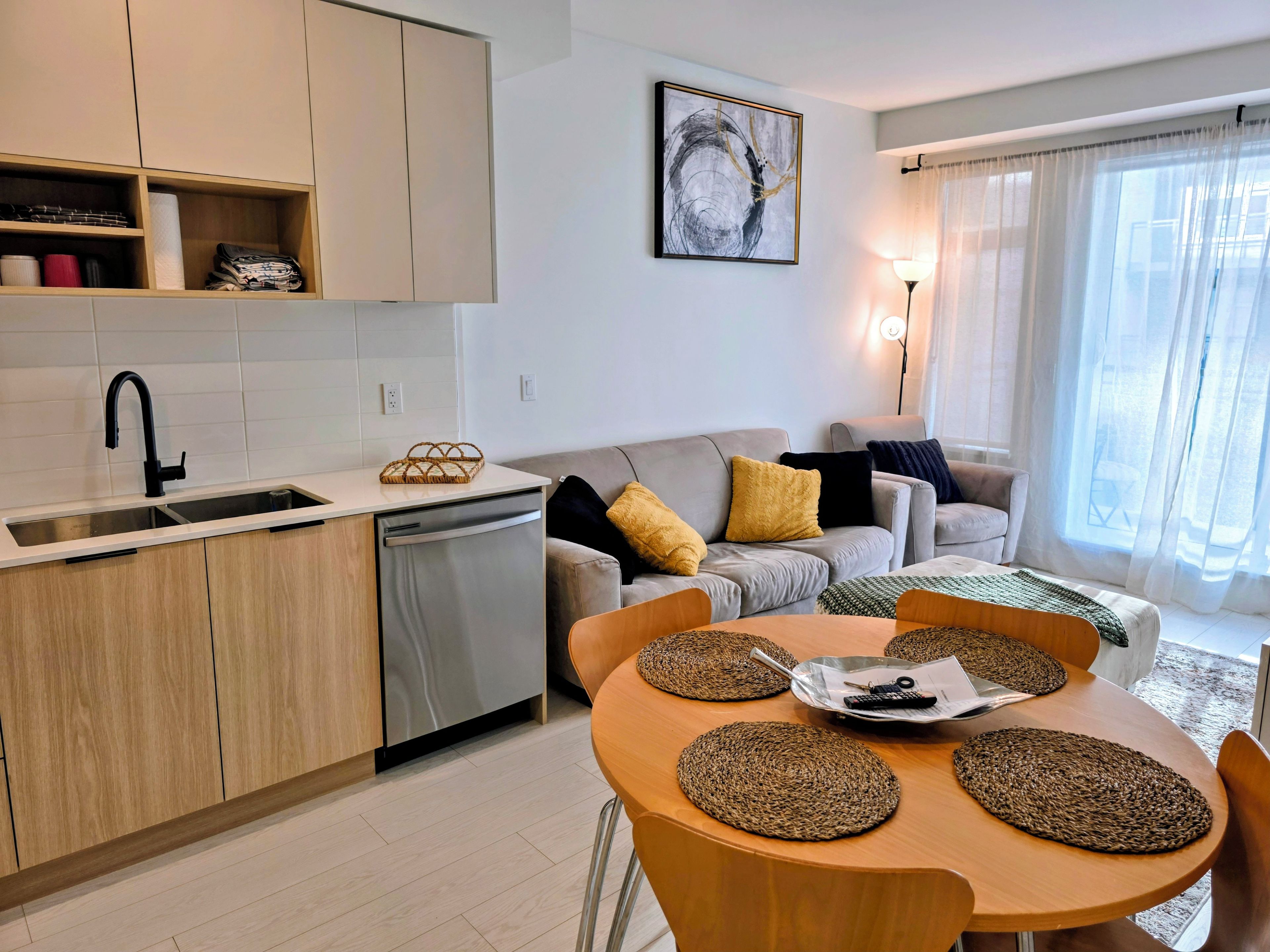 A modern kitchen with a dishwasher, a dining area with a round table and chairs, and a living room with a sofa and a lamp.