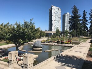 A park with a pond, stone benches, and a statue. In the background, there are tall buildings and trees. - Central Surrey Gem – Steps from SkyTrain, Dining & Groceries (Surrey)