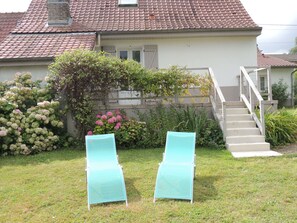 Une maison avec un toit recouvert de tuiles rouges, un mur en pierre et un escalier menant à un balcon. On peut voir deux fauteuils de jardin verts dans la cour.