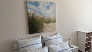 A bedroom with a bed, pillows, a nightstand, a lamp, and a wall-mounted painting of a beach scene. - 300m to the fine Baltic Sea beach - Comfort vacation apartment Seemöwe Scharbeutz (Scharbeutz)