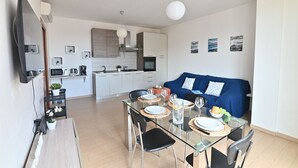 Een moderne keuken met witte kastjes, een magnetron en een koffieapparaat. Er is een eethoek met een glazen tafel voor twee personen, en een blauwe bank in de woonkamer.