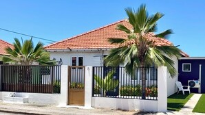 Una casa con techo de tejas rojas, entrada protegida por una verja, un pequeño porche y un edificio azul contiguo.