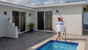 Un espace aménagé au bord de la piscine, avec une piscine, un bâtiment blanc dont les portes sont numérotées et un pot de fleurs.