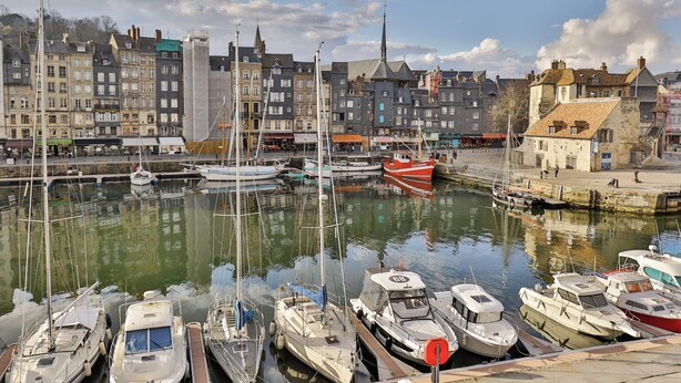 A marina with various boats docked, a waterfront promenade, and historic buildings in the background.
