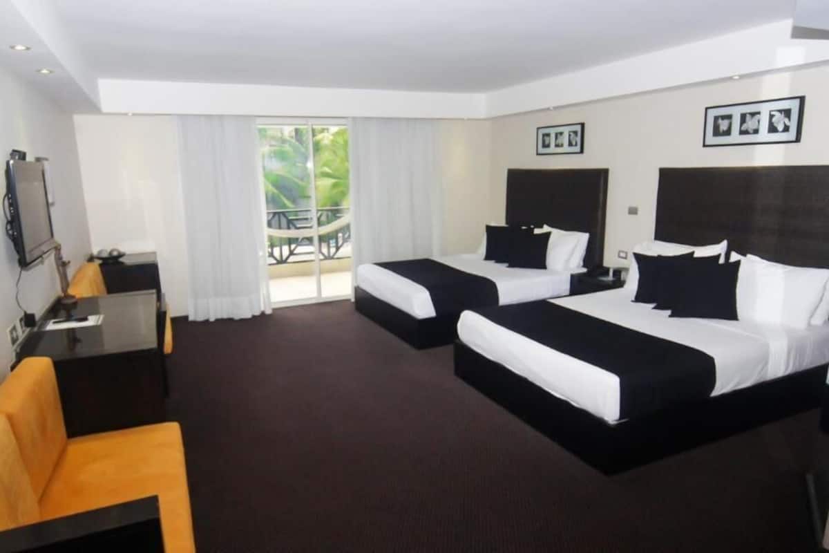 Ein Hotelzimmer mit zwei Betten, einem Schreibtisch, einem Stuhl und einem Balkon mit Blick auf Grünflächen.
