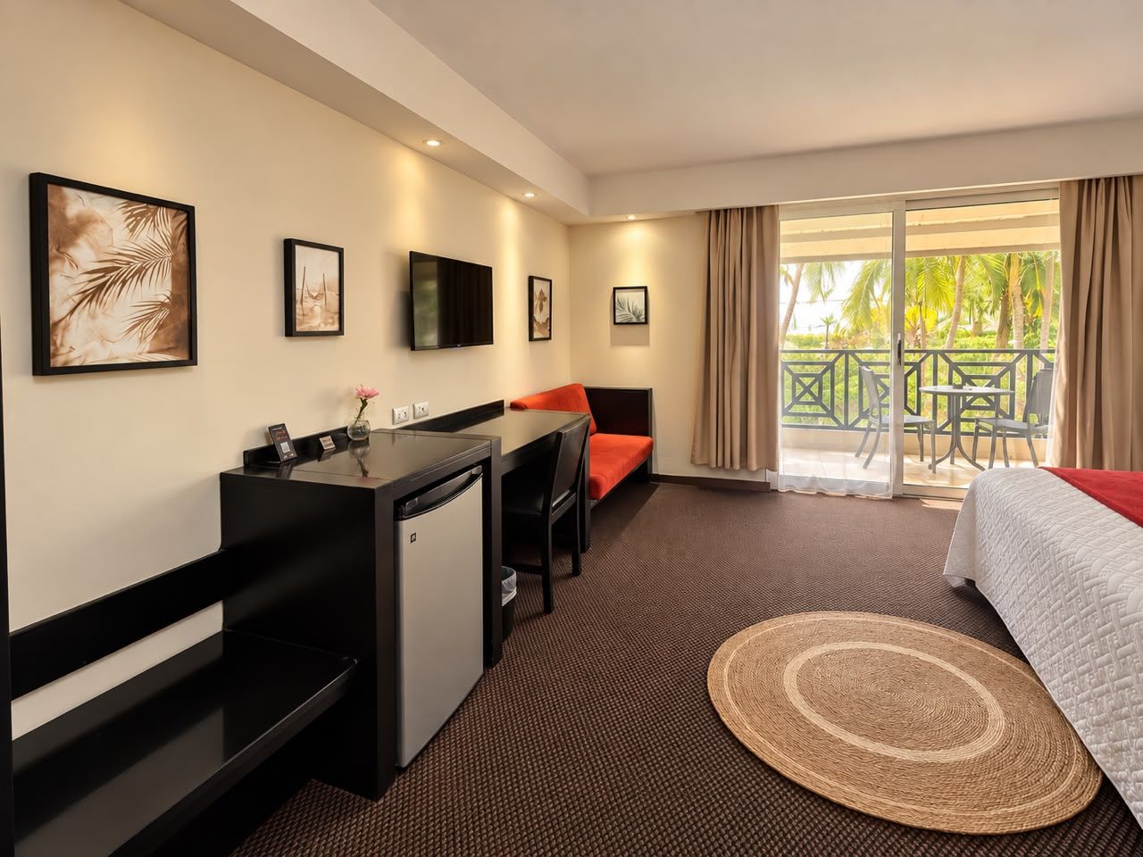 Habitación de hotel con cama, escritorio, silla, una kitchenette pequeña y un balcón con vistas a palmeras.