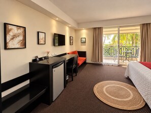 A hotel room with a bed, desk, and chair, a small kitchenette, and a balcony with a view of palm trees.
