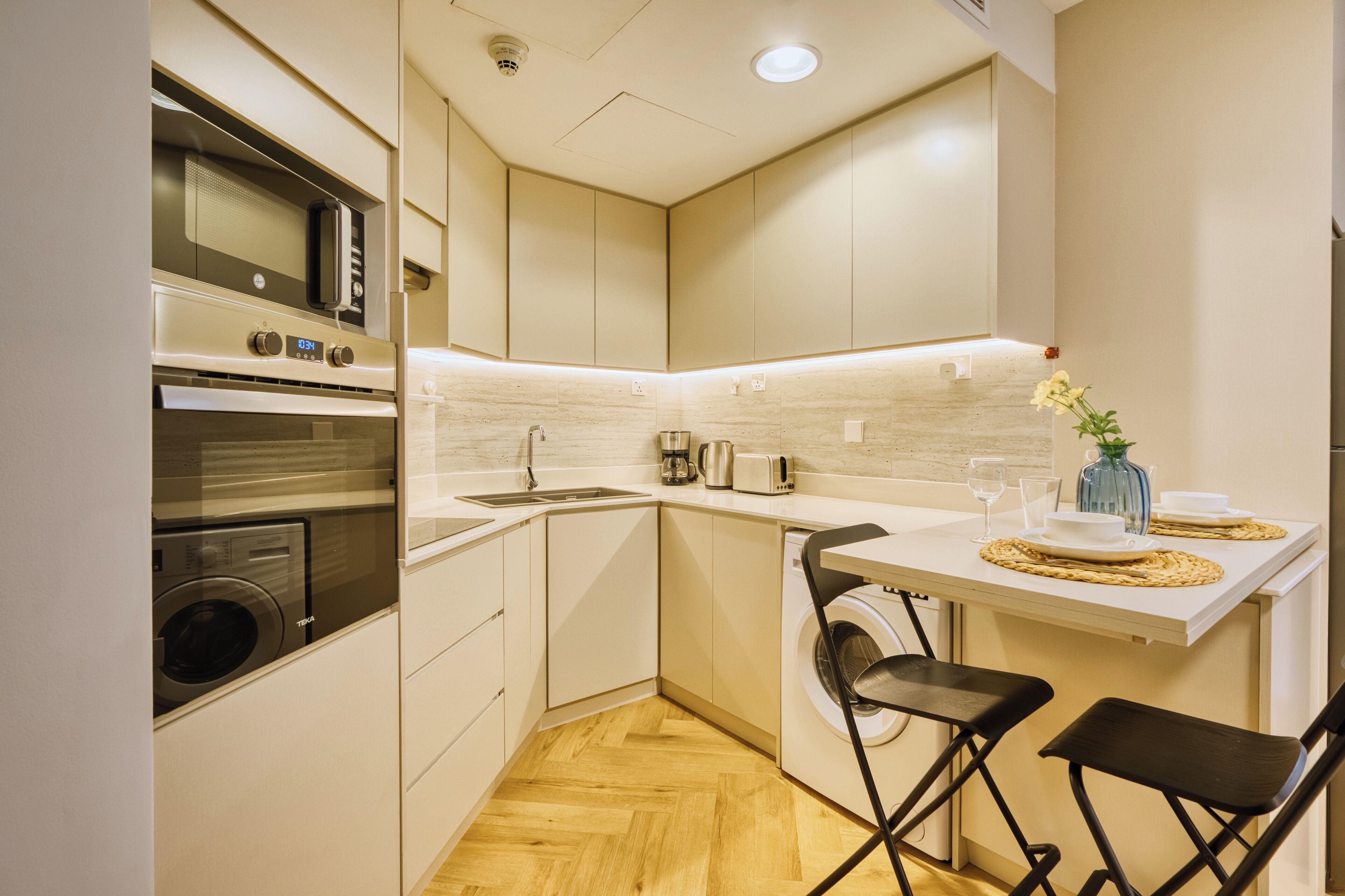 Een compacte keuken met een inbouwoven, magnetron en wasmachine, een klein eethoekje met twee stoelen en een wit aanrecht.