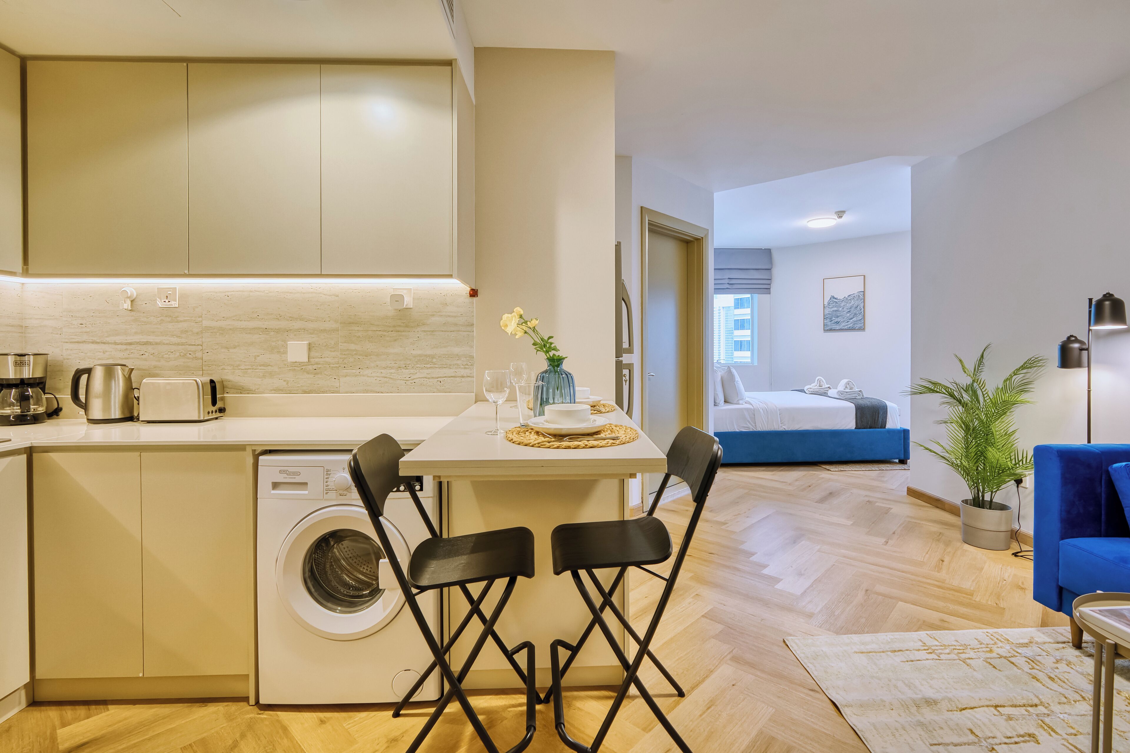 Ett kompakt bostadsutrymme med ett litet kök, en tvättmaskin, en matplats med stolar, en säng, en krukväxt och en blå soffa.