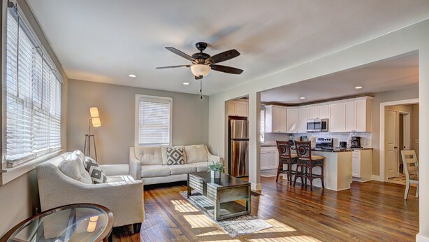A spacious living room with a sofa, coffee table, and a glass-top table. A ceiling fan and a kitchen with white cabinets and a microwave are visible in the background. - Spacious 4-bedroom house in tranquil Decatur with AC (Decatur)