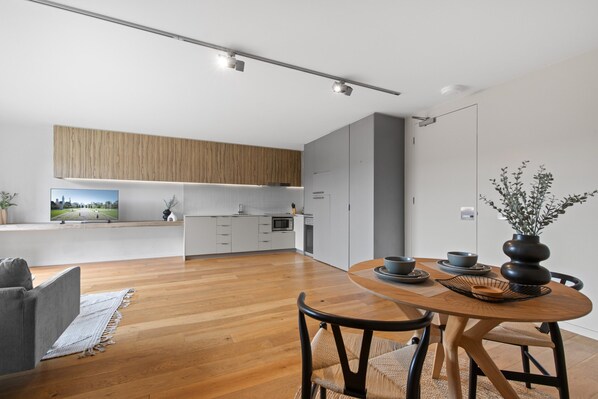 A modern kitchen with wooden cabinets, a dining area with a round table and chairs, and a view of a park through a large window. - Lovely 1-bed With Balcony in St Kilda's Heart (Melbourne)