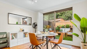 Un comedor con una mesa redonda, sillas naranjas, un armario blanco con libros y un ventanal con vistas a un jardín y una casa.