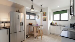 Una cocina moderna con un refrigerador de acero inoxidable, taburetes de bar de madera, una pequeña mesa de comedor y un ventanal con persiana verde.