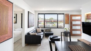 Una sala de estar moderna con un sofá, una mesa de comedor y un televisor. Amplias ventanas ofrecen vistas a edificios cercanos.