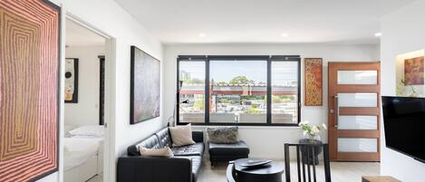 A modern living room with a sofa, a dining table, and a TV. There are large windows with a view of buildings outside.
