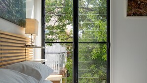 Un dormitorio con cama, mesita de noche, lámpara y un ventanal con vistas al verdor.